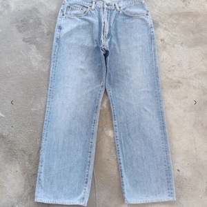 May include: Light blue denim jeans with a classic five-pocket design. The jeans have a straight leg cut and a button and zipper closure. The denim has a faded appearance, suggesting a worn-in look. The jeans are laid flat on a concrete surface.