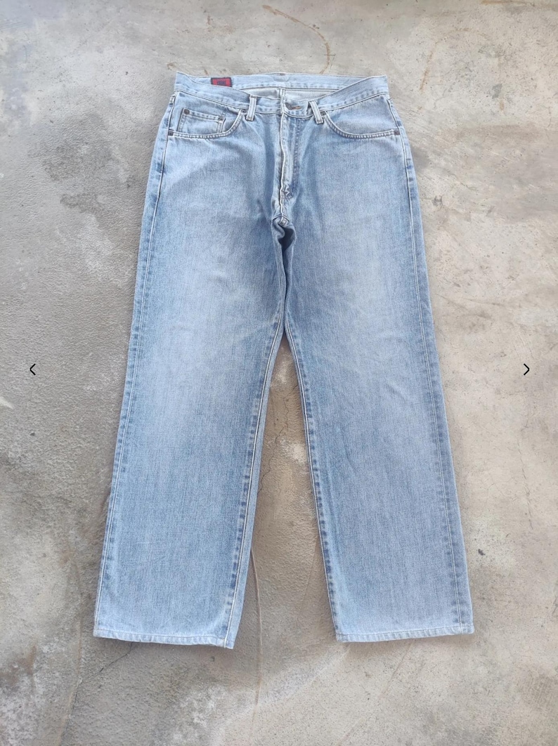 May include: Light blue denim jeans with a classic five-pocket design. The jeans have a straight leg cut and a button and zipper closure. The denim has a faded appearance, suggesting a worn-in look. The jeans are laid flat on a concrete surface.