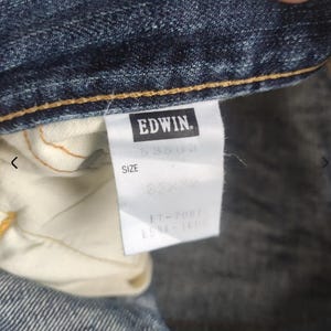 Puede incluir: Primer plano de unos vaqueros azules con una etiqueta blanca. La etiqueta presenta la marca "EDWIN" en un rect&aacute;ngulo negro. La etiqueta tambi&eacute;n muestra la talla "32x32" y otra informaci&oacute;n del producto.