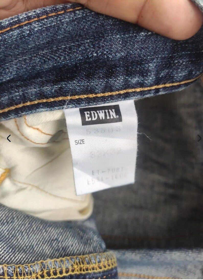 Puede incluir: Primer plano de unos vaqueros azules con una etiqueta blanca. La etiqueta presenta la marca "EDWIN" en un rect&aacute;ngulo negro. La etiqueta tambi&eacute;n muestra la talla "32x32" y otra informaci&oacute;n del producto.