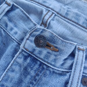 May include: Close-up of light blue denim jeans. The jeans feature a button closure with the brand name "EDWIN" embossed on the button. The stitching is a contrasting brown color, and the fabric shows signs of wear.