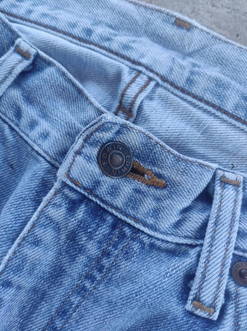 May include: Close-up of light blue denim jeans. The jeans feature a button closure with the brand name "EDWIN" embossed on the button. The stitching is a contrasting brown color, and the fabric shows signs of wear.