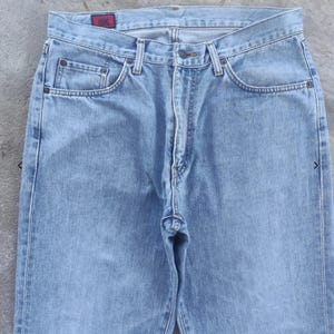 May include: Light blue denim jeans with a classic five-pocket design. The jeans have a button and zipper closure, belt loops, and a small red tag on the back pocket. The denim appears to be pre-washed for a faded look.