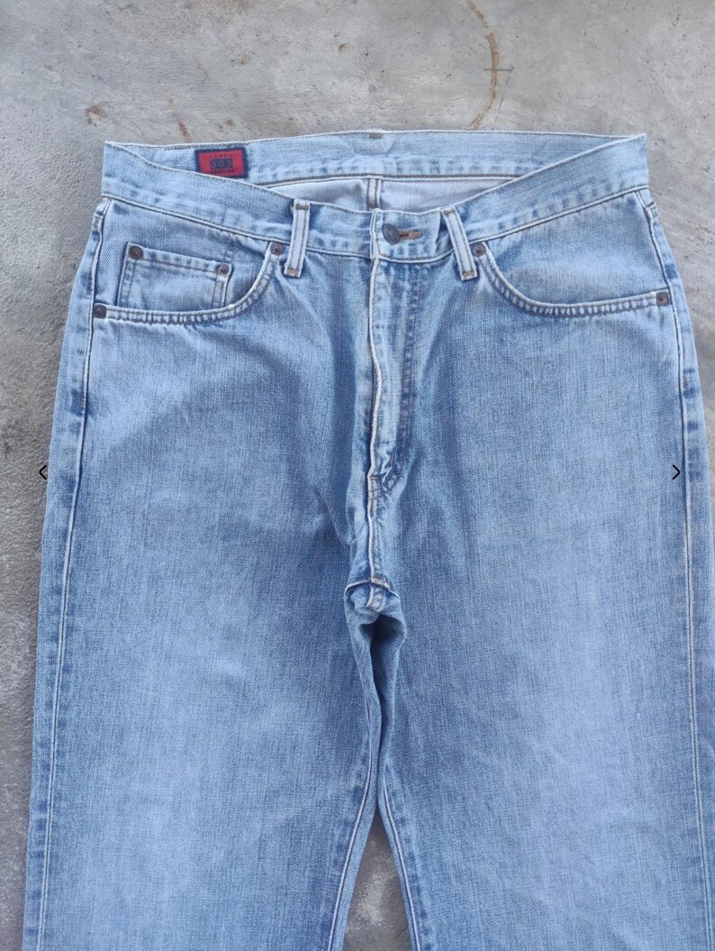 May include: Light blue denim jeans with a classic five-pocket design. The jeans have a button and zipper closure, belt loops, and a small red tag on the back pocket. The denim appears to be pre-washed for a faded look.