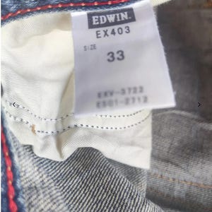 Puede incluir: Primer plano de una prenda de mezclilla azul con una etiqueta blanca. La etiqueta dice "EDWIN. EX403 SIZE 33" y texto adicional. Se aprecia costura roja en el borde de la mezclilla.