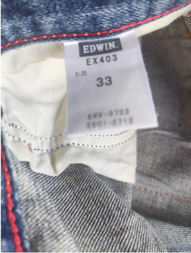 Puede incluir: Primer plano de una prenda de mezclilla azul con una etiqueta blanca. La etiqueta dice "EDWIN. EX403 SIZE 33" y texto adicional. Se aprecia costura roja en el borde de la mezclilla.
