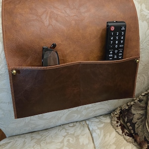 May include: A brown leather couch arm organizer with two pockets. One pocket holds a remote control and the other holds a pair of glasses. The organizer is designed to fit over the arm of a couch.