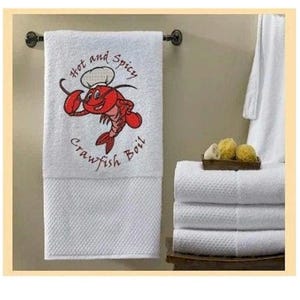 May include: White bath towel with a textured bottom section, hanging on a dark metal rod. The towel features an embroidered cartoon crawfish chef with the text "Hot and Spicy Crawfish Boil" in red and black.