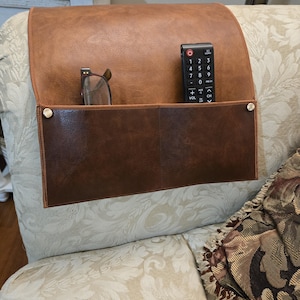 May include: A brown faux leather couch armrest organizer with two pockets. The top pocket holds a remote control and the bottom pocket holds a pair of eyeglasses. The organizer is attached to the couch with two gold-colored snaps.