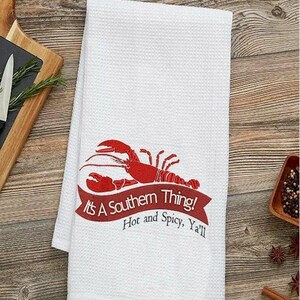May include: White kitchen towel featuring a red lobster design and the text "It's A Southern Thing! Hot and Spicy, Ya'll". The towel is folded on a wooden surface.