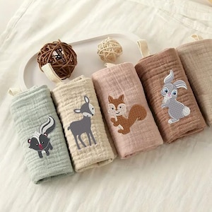 May include: A collection of six rolled washcloths in various pastel colors, each embroidered with a different woodland animal: a skunk, deer, squirrel, and rabbit. Each cloth has a loop for hanging. The washcloths are made of a soft, textured fabric.