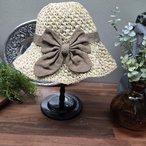 May include: A beige straw hat with a wide brim and a brown bow. The hat is displayed on a black stand. The bow is made of a textured fabric. The background is neutral.