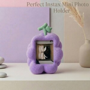 May include: A lilac-coloured photo holder with a distinctive, bubbly design. The holder has a green, leaf-like accent at the top and displays a small photo. The text "Perfect Instax Mini Photo Holder" is printed on the image.