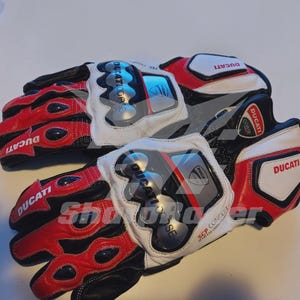 Peut inclure: Paire de gants de moto rouges, blancs et noirs avec le logo Ducati. Les gants ont un rembourrage de protection sur les articulations et la paume. Le texte "SCP Concept" est visible sur les gants.