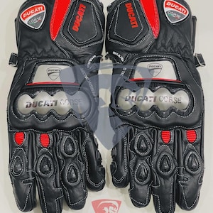 Peut inclure: Paire de gants de moto en cuir noir avec des détails rouges et argentés. Les gants présentent le logo et le texte "Ducati Corse". Des coutures blanches soulignent le design. Les gants ont une protection des articulations et une sangle de poignet. Le logo "Pythons Racing Gear" est visible.