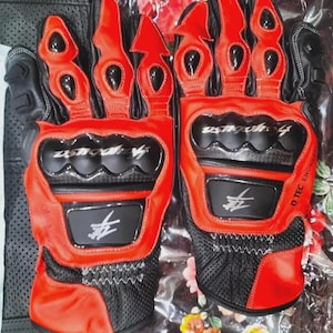 Può includere: Paio di guanti da moto rossi e neri. I guanti presentano pelle rossa e tessuto a rete nero. Hanno pannelli protettivi neri sulle nocche e sui palmi. I guanti mostrano il testo "Ygmouse" e un simbolo.