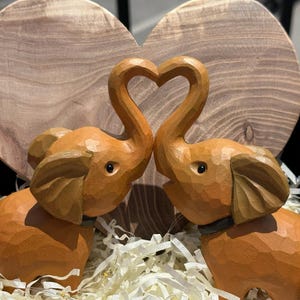 May include: Two wooden elephant figurines are facing each other, their trunks forming a heart shape. The elephants are a light brown color and have detailed carvings. They are sitting on a bed of white shredded paper inside a red box.