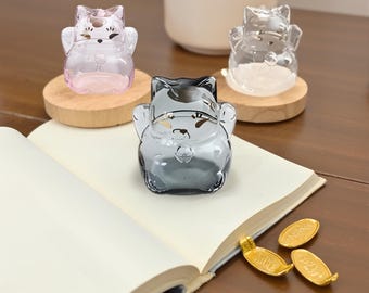 May include: Three glass cat figurines in pink, clear, and gray, each on a small wooden base. The figurines are in a welcoming pose. The scene includes an open book and gold-colored coins.