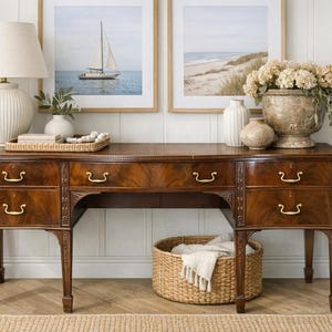 May include: A dark wood desk with six drawers and brass handles. A woven basket with a blanket sits below the desk. The desk is decorated with a lamp, vases, and framed artwork. The room has a neutral color palette.