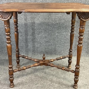 May include: A brown, antique side table with a scalloped top and decorative carved fan details on each end. The table has turned legs and a lower shelf for added stability. The table is approximately 30 inches long.