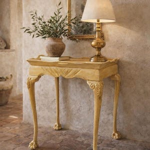 May include: A light yellow, antique-style side table with carved details and claw feet. A gold-colored lamp with a beige lampshade sits on the table, along with a vase of greenery and a decorative mirror.