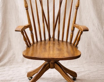 May include: A wooden swivel desk chair with a spindle back and armrests. The chair has a round seat and is supported by a base with four legs and small caster wheels. The wood has a warm, medium brown tone.
