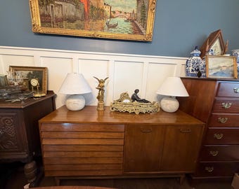 Puede incluir: Un aparador de madera con una sección izquierda de puertas con listones horizontales y una sección derecha con dos puertas lisas. Dos lámparas blancas, una figura de ángel dorada y un reloj decorativo descansan sobre el mueble.