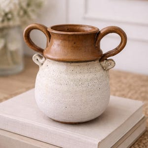 May include: A ceramic vase with a rounded body and two handles. The top portion is brown, while the lower part is off-white. The vase is sitting on top of two books with a neutral color.