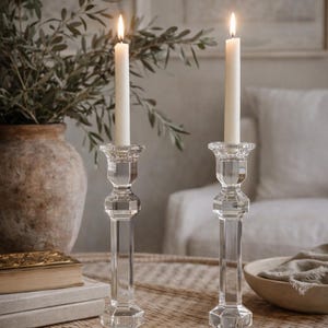 May include: Two clear glass candle holders with lit white candles. The candle holders have a faceted design and sit on a woven table. A vase with greenery and a stack of books are in the background.