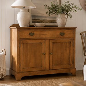May include: A wooden cabinet with two doors and a drawer, featuring brass hardware. A lamp with a cream-colored shade and a vase with greenery sit on top. A woven basket and a chair are also in the image.