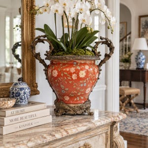 May include: A vibrant orange ceramic urn featuring a delicate floral pattern and ornate bronze-toned metal handles and base. The urn is filled with white orchid flowers and sits atop a stack of three books titled At Home, More Beautiful, and The Kinfolk Home.