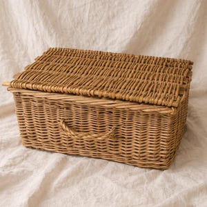 Puede incluir: Una cesta rectangular de mimbre con tapa con bisagras y asa. La cesta est&aacute; tejida con mimbre marr&oacute;n claro y se encuentra sobre un fondo de tela blanca. La cesta est&aacute; dise&ntilde;ada para el almacenamiento.
