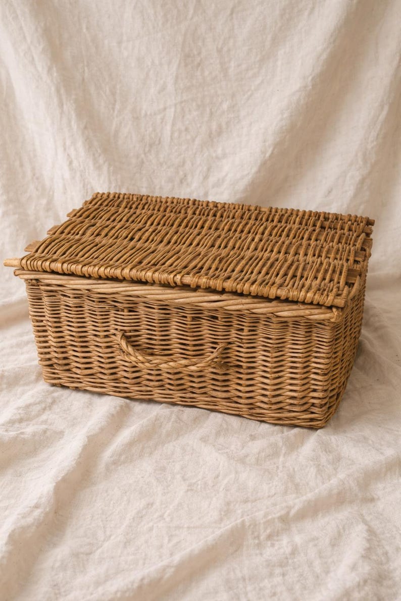 Puede incluir: Una cesta rectangular de mimbre con tapa con bisagras y asa. La cesta est&aacute; tejida con mimbre marr&oacute;n claro y se encuentra sobre un fondo de tela blanca. La cesta est&aacute; dise&ntilde;ada para el almacenamiento.