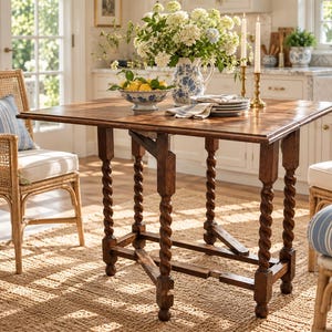 May include: A rectangular wooden drop-leaf table featuring a dark brown finish and four barley twist legs. The table is supported by a central gate-leg mechanism and stands on a woven natural fiber rug, accompanied by two rattan chairs with cushions.
