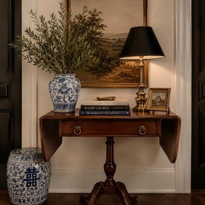 May include: A dark wood table with a drop-leaf design, holding a blue and white floral vase with greenery, books, and a lamp with a black shade. A matching blue and white garden stool sits to the left. A framed landscape painting hangs above.