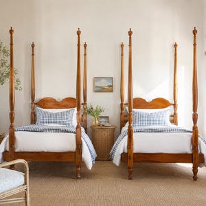 May include: Two twin-sized four-poster beds made of polished brown wood, each featuring a white duvet and a blue and white gingham patterned pillow. The beds have tall, slender posts and arched headboards, positioned symmetrically in the room.