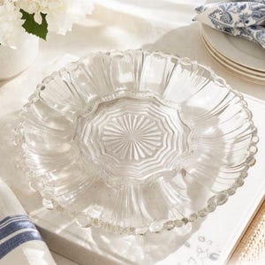 May include: A clear glass serving dish with a scalloped edge and a central starburst design. The dish is round and sits on a white surface, with a white vase of flowers and a stack of plates in the background.