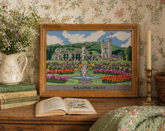 May include: A framed needlepoint of Balmoral Castle, featuring a detailed landscape with a castle, fountain, and colorful flowerbeds. The artwork is displayed on a wooden surface with books, a vase of flowers, and a candle holder.