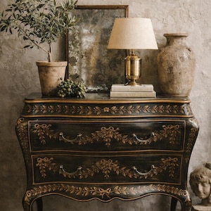 May include: A black antique chest of drawers with gold floral detailing. A small potted olive tree, a mirror, a brass lamp, and a textured vase sit atop the chest. The scene is set against a textured gray wall.