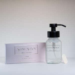 May include: A clear glass bottle with a black pump dispenser, a rectangular box, and a small white bar. The bottle and box have the text "SCENT SI BON". The box is light purple with silver text. The bottle is filled with a clear liquid.