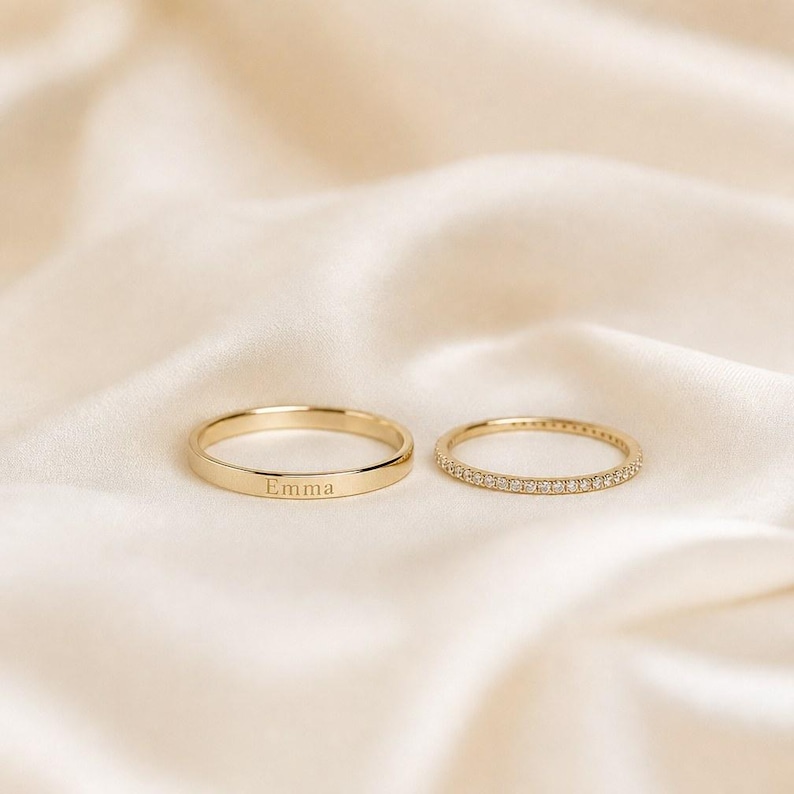May include: Two gold rings. One ring is a smooth, plain band with the name Emma engraved on the outer surface. The second ring is a thin, delicate band featuring a continuous row of small, clear, sparkling stones set along the entire circumference.