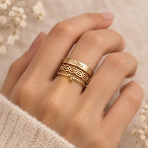 Könnte beinhalten: Ein Set aus drei goldfarbenen Ringen, die übereinander getragen werden. Der oberste Ring ist ein glattes Band mit der Gravur Emma. Der mittlere Ring zeigt ein komplexes, sich wiederholendes Kettenmuster. Der untere Ring ist ein dünner, offener Reifen.