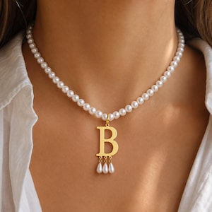 May include: A necklace featuring a strand of white round pearls and a gold-toned letter B pendant. The pendant is adorned with three small, dangling white teardrop-shaped pearls attached to the bottom of the letter with gold-toned jump rings.