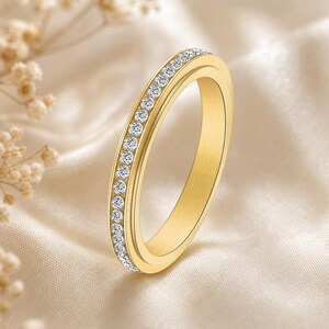 May include: A gold-toned band ring featuring a single row of small, clear, round-cut stones embedded along the centre. The metal surface has a smooth, polished finish, and the design maintains a consistent width throughout the entire circular band.