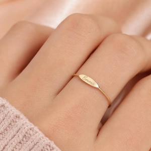 Peut inclure: Une fine bague en or dotée d'une petite pièce centrale plate de forme ovale, gravée du prénom Emma dans une police d'écriture cursive. L'anneau est fin et poli, conservant une teinte dorée métallique uniforme sur toute la pièce.