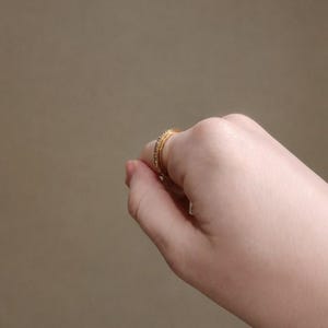 May include: A gold-toned ring featuring a thin band with a row of small, clear, sparkling stones embedded along the outer surface. The ring is shown positioned on a finger, displaying its circular shape and metallic finish in a side profile view.