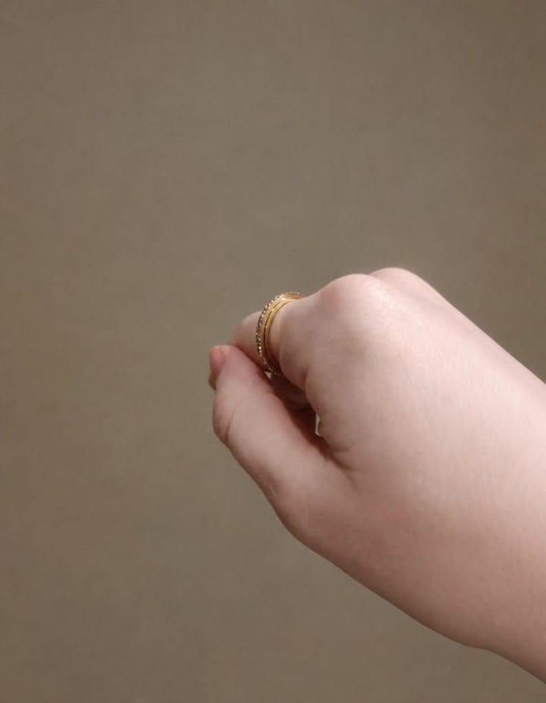 May include: A gold-toned ring featuring a thin band with a row of small, clear, sparkling stones embedded along the outer surface. The ring is shown positioned on a finger, displaying its circular shape and metallic finish in a side profile view.
