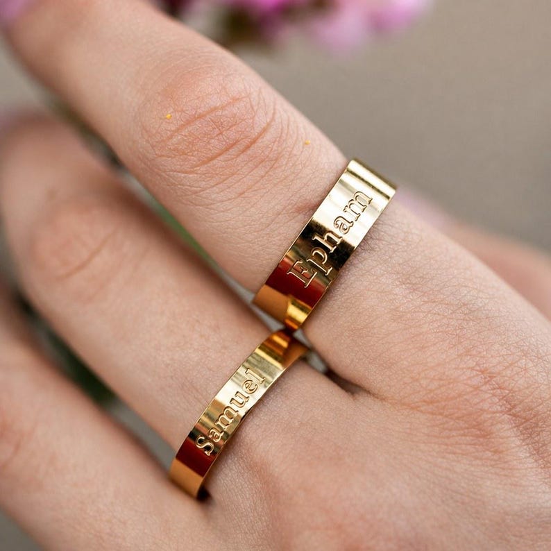May include: Two polished gold-tone metal bands featuring engraved text. The upper ring displays the name Ephram in a serif font, while the lower ring features the name samuel in a lowercase sans-serif font. Both rings have a flat, smooth surface.