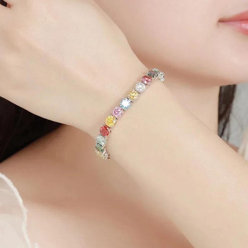 May include: A silver bracelet adorned with a series of colorful, round gemstones. The gemstones are arranged in a row, showcasing a spectrum of colors including red, yellow, green, and blue. The bracelet is worn on a wrist.