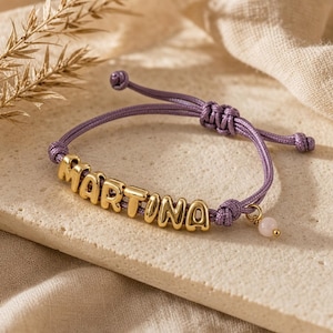 Op de afbeelding: Een paarse koordarmband met goudkleurige metalen letterkralen die de naam MARTINA vormen. De verstelbare armband heeft een kleine, ronde, lichtroze kraal als bedel die aan de zijkant bij de knoopsluiting hangt.